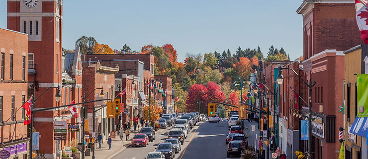 Downtown Bracebridge: downtown buildings framing the picture, autumn coloured trees in the background, a street full of cars and pedestrians shopping.
