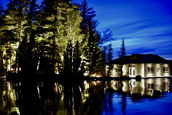 Outdoor lighting project in Ontario by Nightscapes: a well-lit cottage on the lake. A very blue sky and lots of trees nearby. There is a reflection on the water.